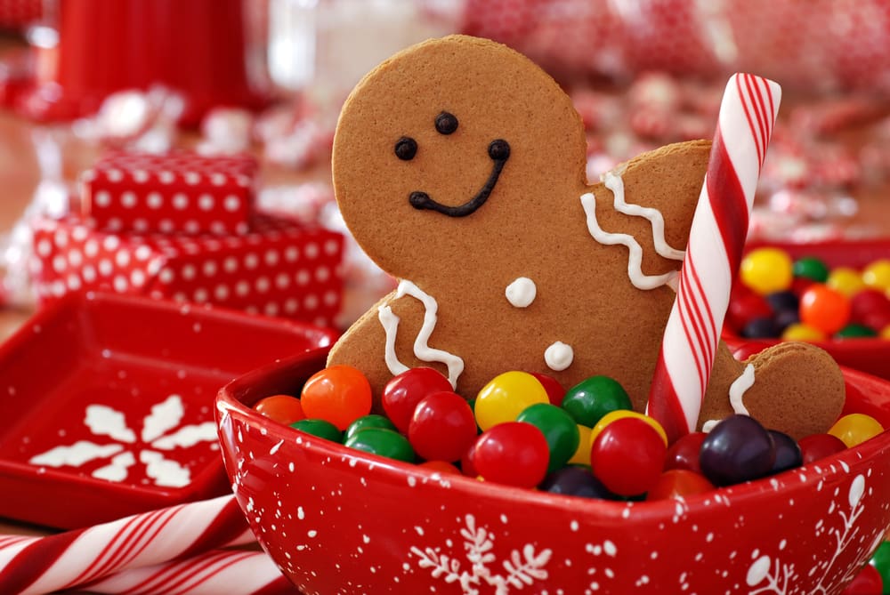 Smiling gingerbread man holding a peppermint stick in a red snowflake dish with colourful sweets. Wrapped gifts, sweets and red dishes in the background