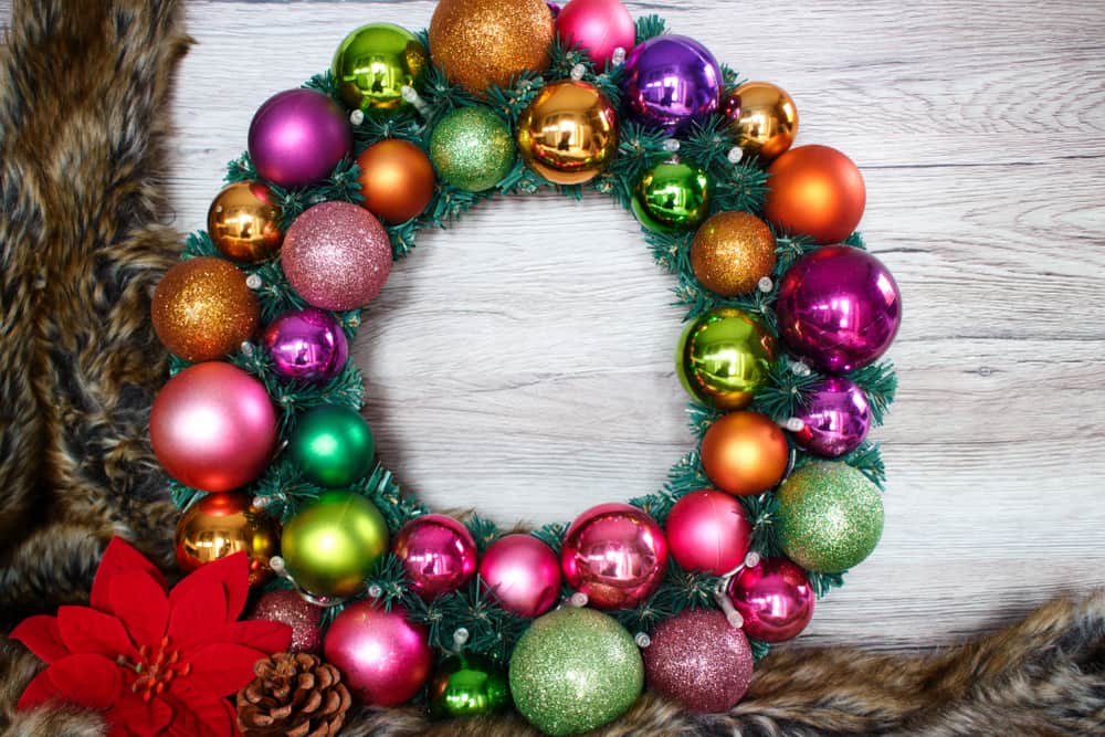 Multi-coloured bauble wreath with lights sat on a fur coat with a wooden background and a red poinsettia flower in the bottom left hand corner