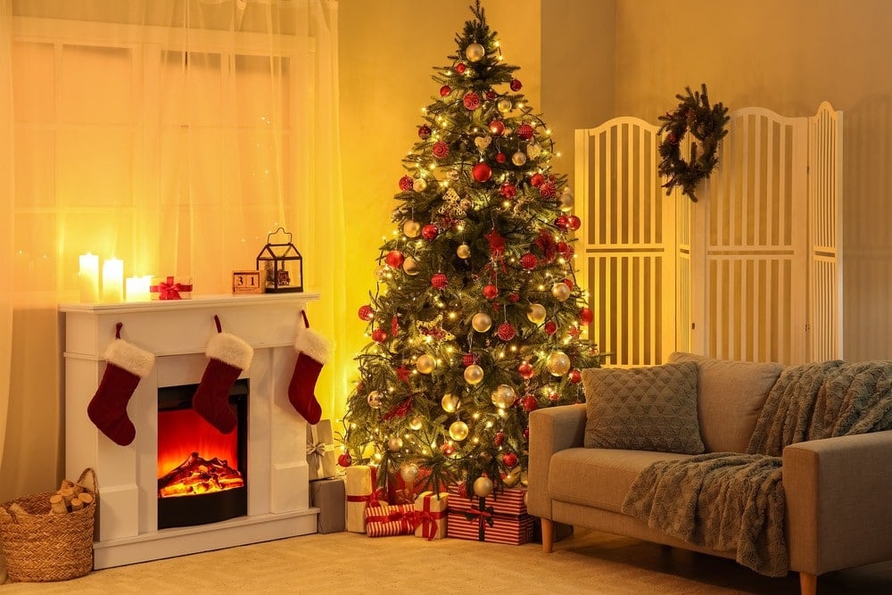 Living room scene with a Christmas tree. To the left is a fireplace with three stockings and some ornaments and to the right is a grey sofa and a white partition with a wreath hanging from it